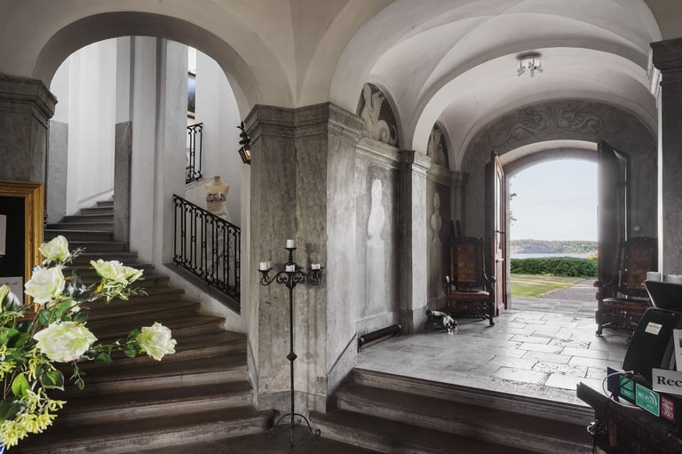 Grand entryway with arched ceilings, wooden staircase on left, open doorway on right with countryside view, and white hydrangea flowers