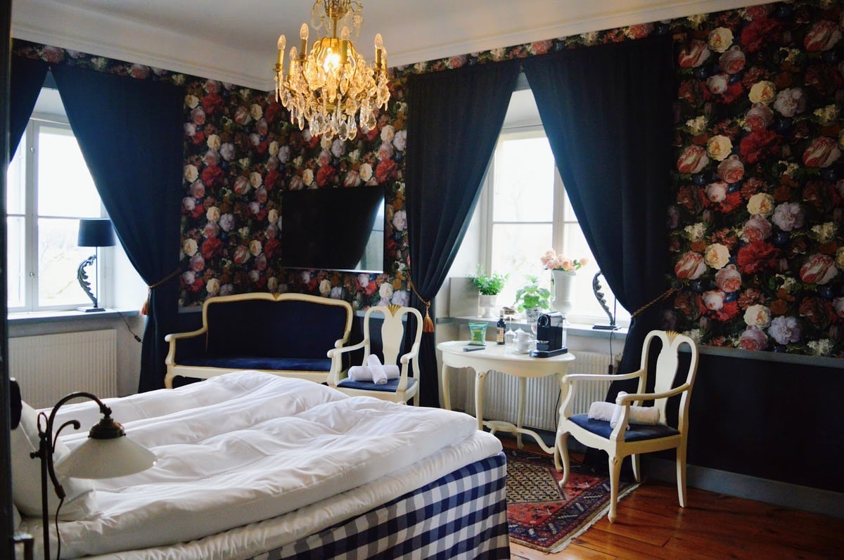 Elegant bedroom with dark floral wallpaper, gold chandelier, navy curtains, white bedding, and ornate furniture