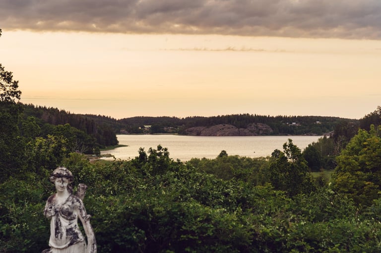 Stone statue in foreground overlooking forested landscape and calm lake under cloudy sky at dusk