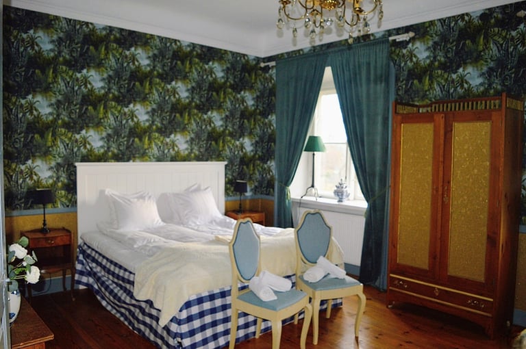 Elegant bedroom with floral wallpaper, white bed with blue gingham skirt, wooden wardrobe, teal curtains, and period furnishings