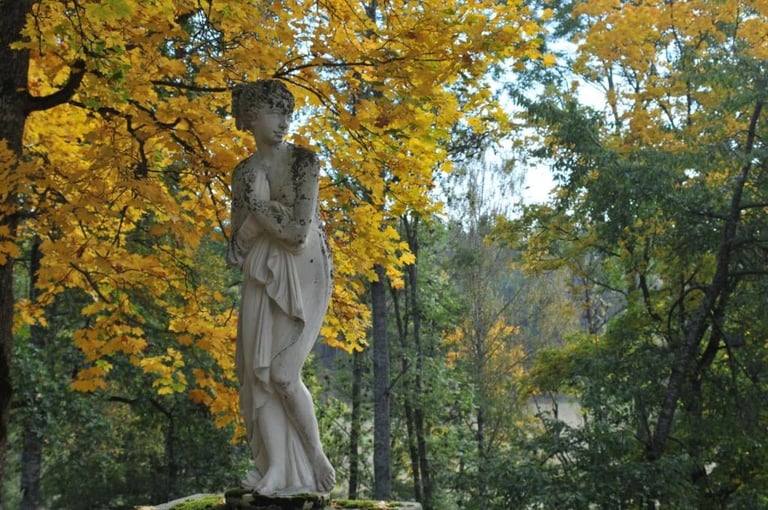 White marble statue of a robed female figure in a park surrounded by golden autumn foliage and green trees