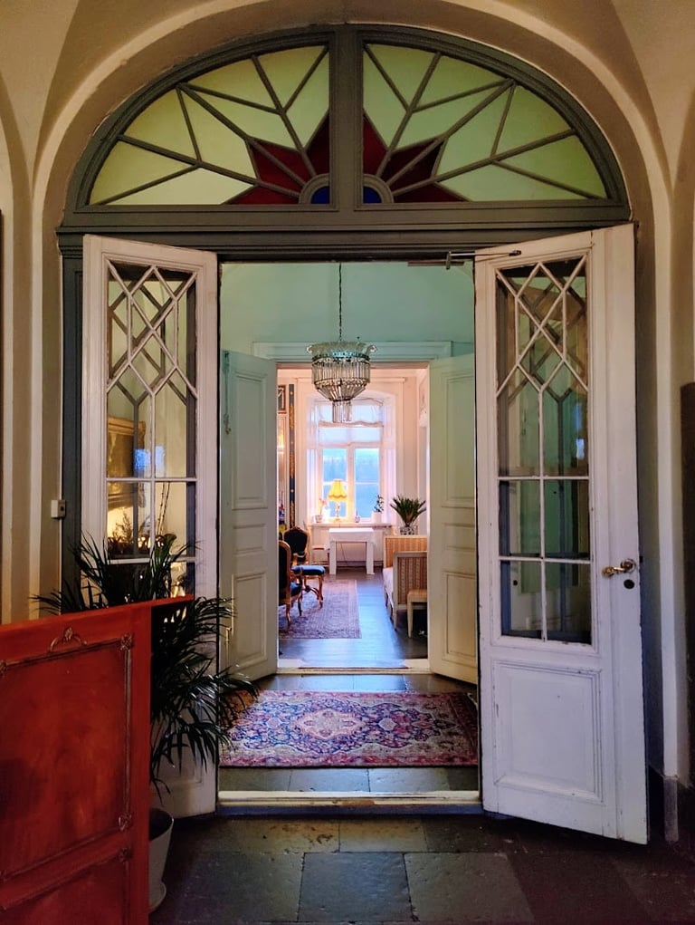 Elegant double doors with stained glass transom open into a bright interior hallway with a chandelier and arched entryway