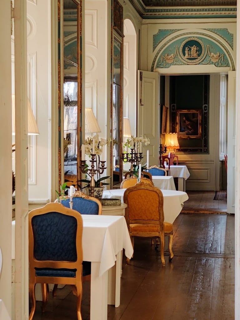 Elegant formal dining room with cream-colored walls, gold-framed mirrors, wooden chairs with blue upholstery, white tablecloths, and ornate classical decor