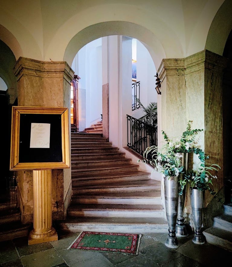 Grand classical staircase with arched corridor, wooden steps, ornate frame on wall, potted plants, and wrought iron railings