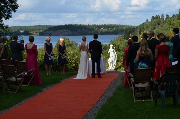 Outdoor lakeside wedding ceremony with bride and groom on red aisle, guests seated on both sides overlooking forested lake