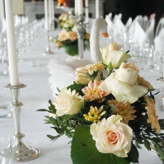 Elegant floral table centerpiece with white and peach roses, yellow daisies, and greenery in a green vase next to a silver candlestick