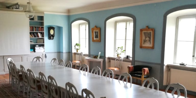 Large dining room with long white table, wooden chairs, blue-green walls, arched windows, shelving, and framed artwork