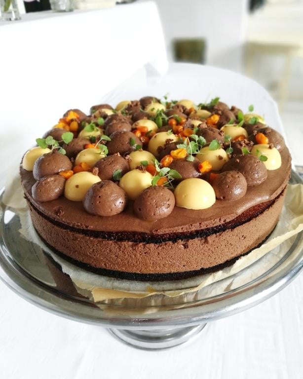 Chocolate layer cake decorated with chocolate truffles, gold candies, and green garnish on a glass cake stand
