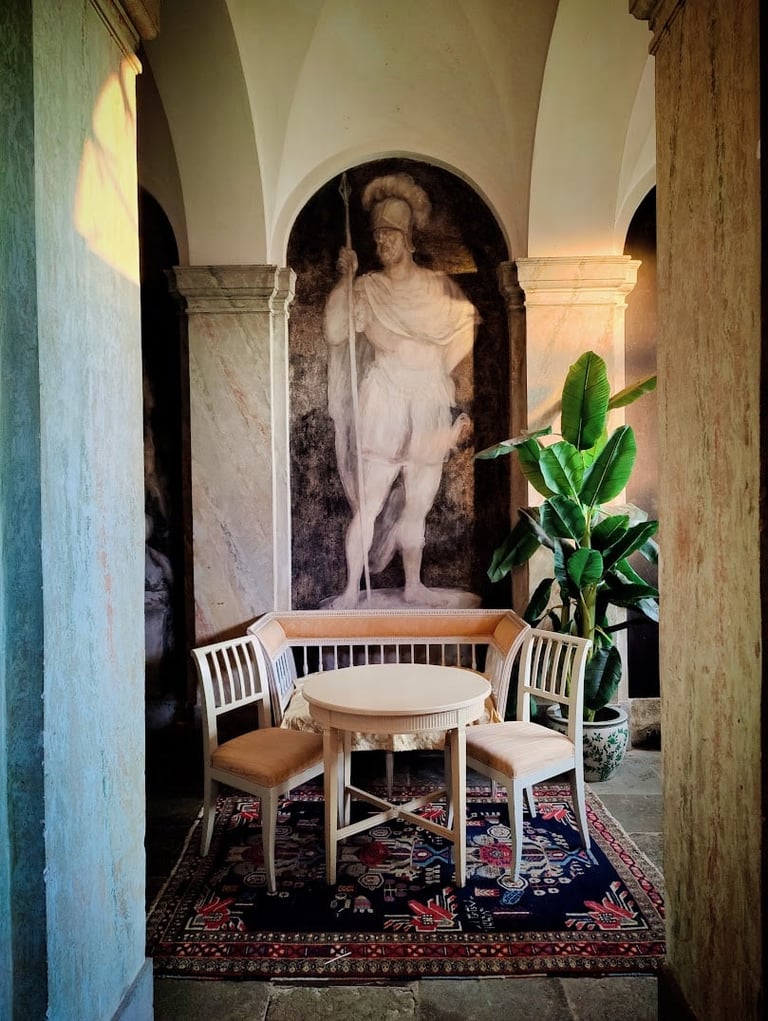 Arched alcove with classical nude statue, round table, two white chairs, and potted green plant on patterned rug