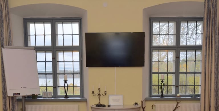 A yellow living room with a flat-screen TV mounted on the wall between two large windows with gray frames overlooking outdoors.