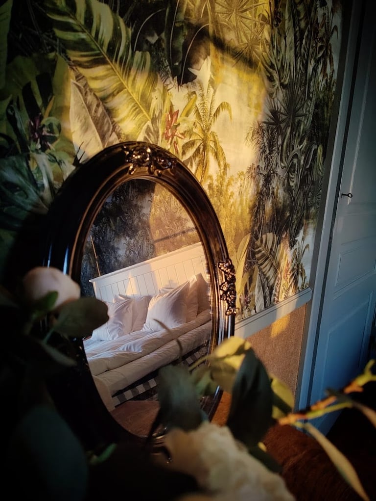 Ornate oval mirror reflecting a bed, set against a botanical wallpaper with golden lighting and plant stems in foreground
