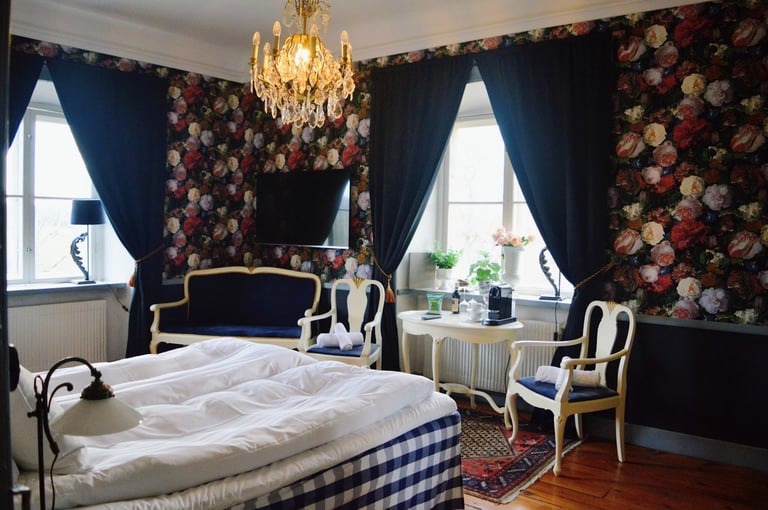 Luxurious bedroom with navy floral wallpaper, gold chandelier, dark curtains, striped bed, ornate furniture, and two large windows
