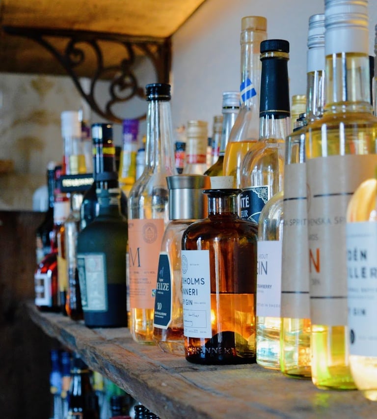 Collection of spirits and liquor bottles arranged on a wooden shelf with varying colors and labels visible