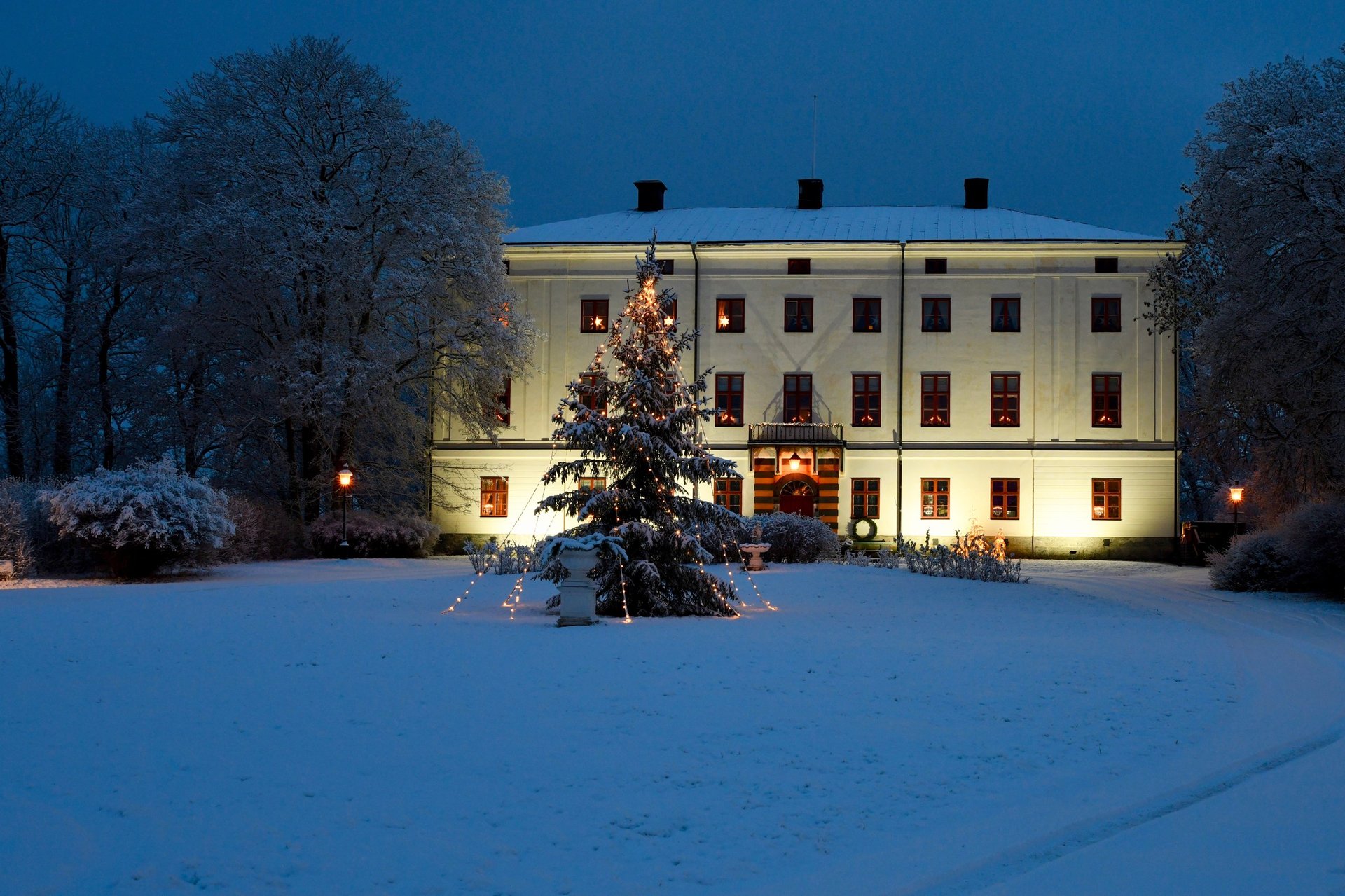 Illuminated white manor house surrounded by snow-covered trees and grounds at dusk with decorated evergreen in foreground