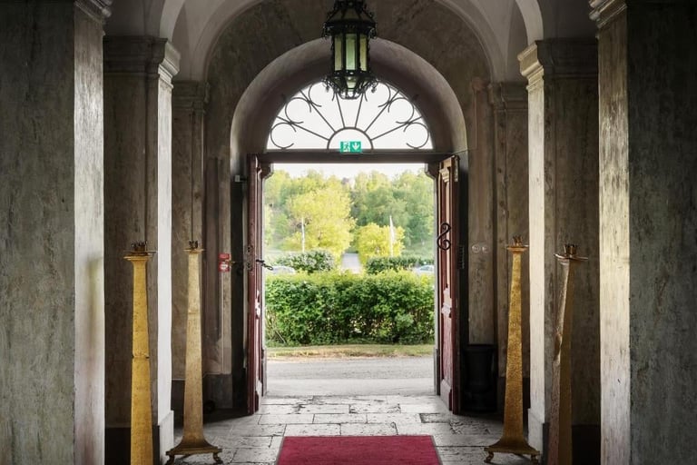 Historic arched entryway with wooden doors opening to a garden, featuring stone columns, a red carpet, and a fanlight window above