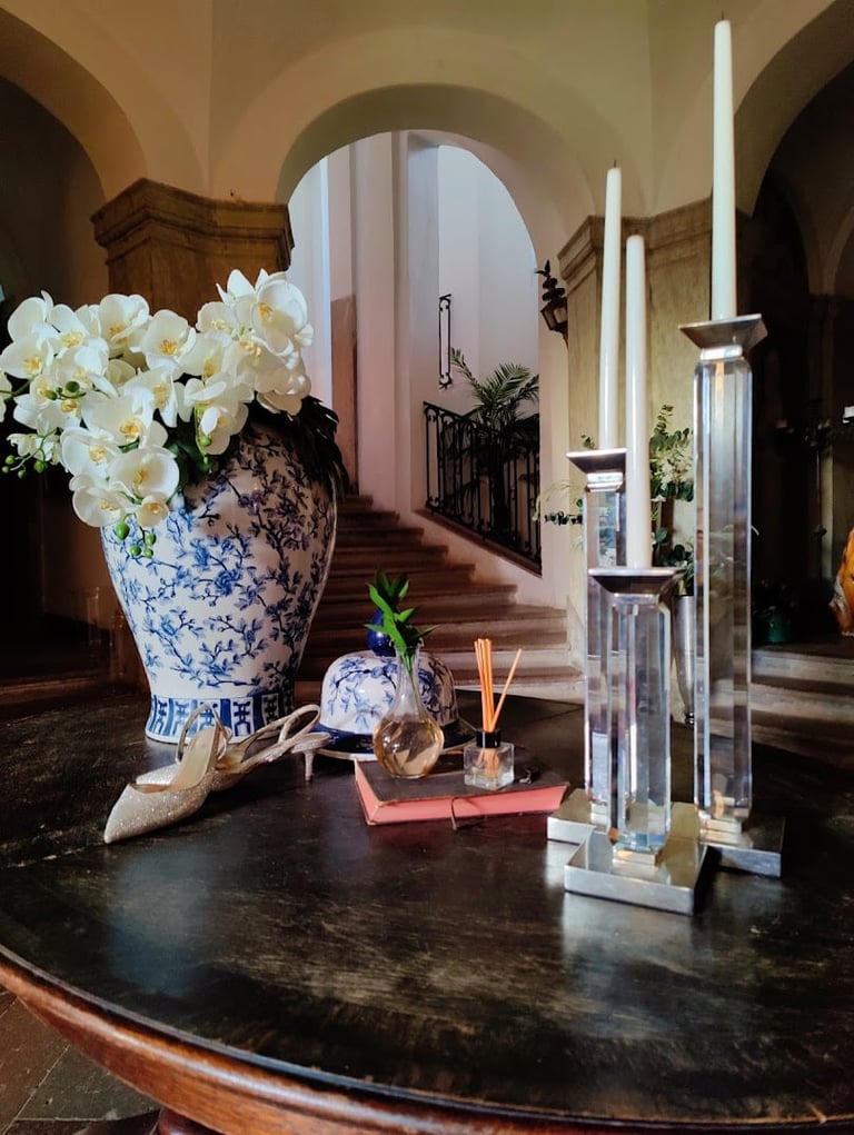 Elegant entryway table with white flowers in blue and white vase, tall candles, and decorative objects, arched hallway in background