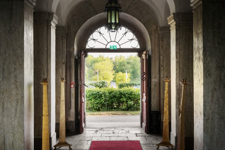Arched stone corridor with columns leading to open doorway, red mat on floor, green garden visible outside