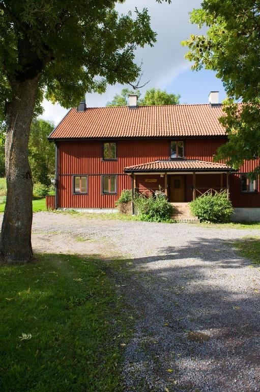 Red corrugated metal farmhouse with terracotta roof surrounded by lush green trees and gravel driveway