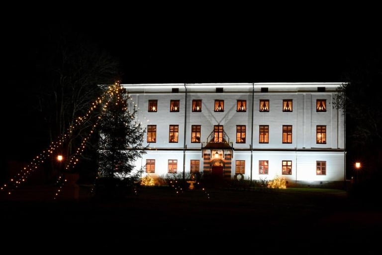 Nighttime view of a white-painted institutional building with illuminated windows, flanked by string lights and trees