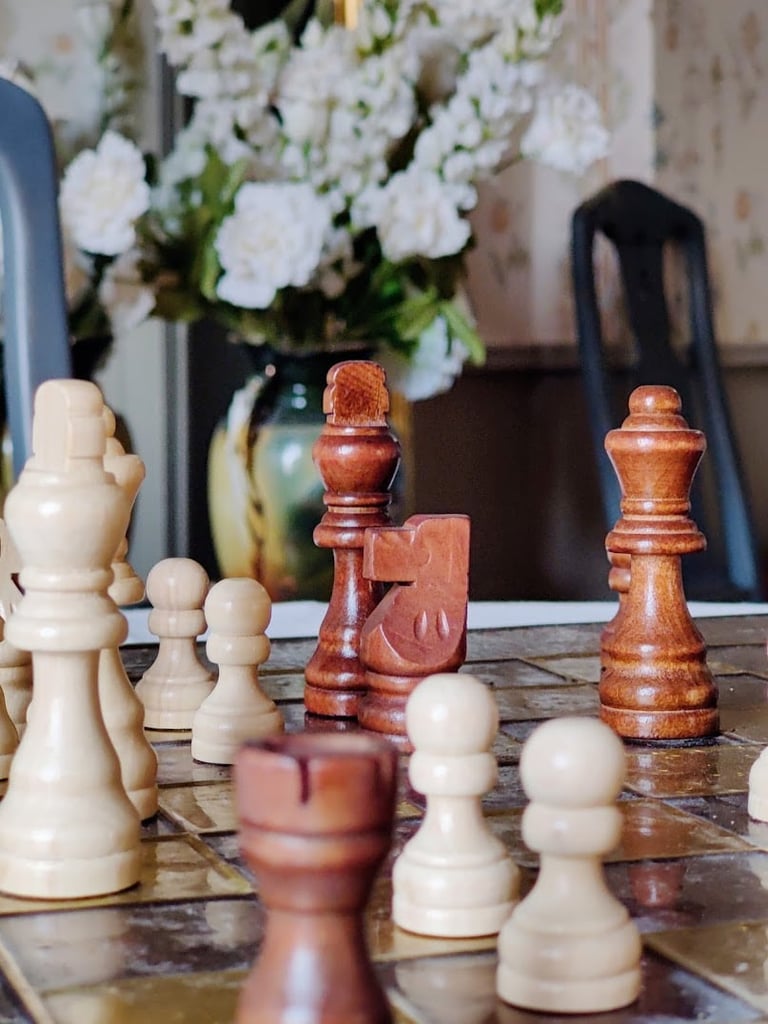 Chess pieces arranged on board with white flowers in vase and black chairs in background