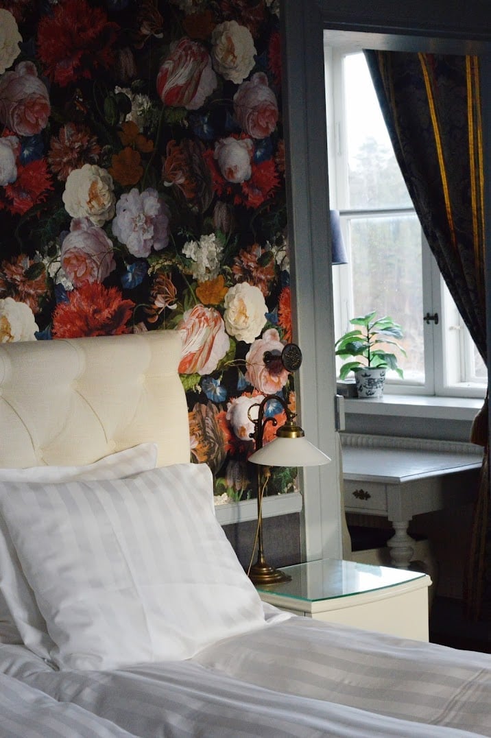 Elegant bedroom with white bedding, floral wallpaper featuring red and pink roses behind headboard, window with black curtains, and nightstand with lamp and plant