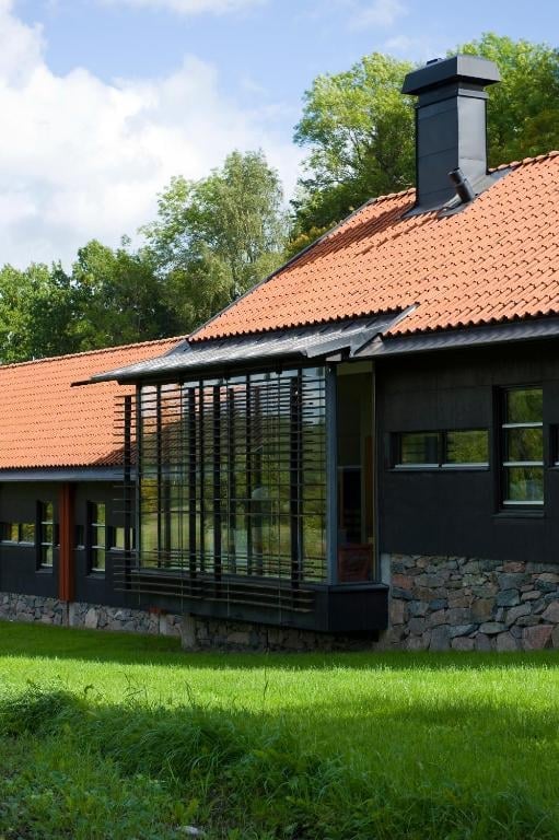 Modern house with terracotta tile roof, black exterior walls, large windows, stone foundation, and surrounded by green lawn and trees
