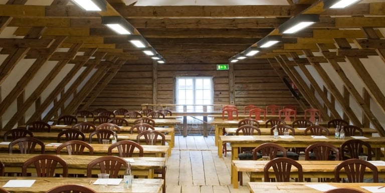 Rustic barn dining hall with wooden beams, tables, chairs, and skylights leading to exit doors