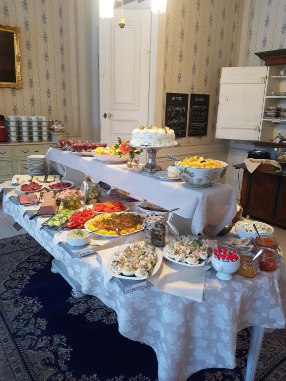 Elegant buffet spread with multiple white tablecloths displaying various dishes, desserts, and appetizers in a formal dining room with vintage wallpaper and furnishings
